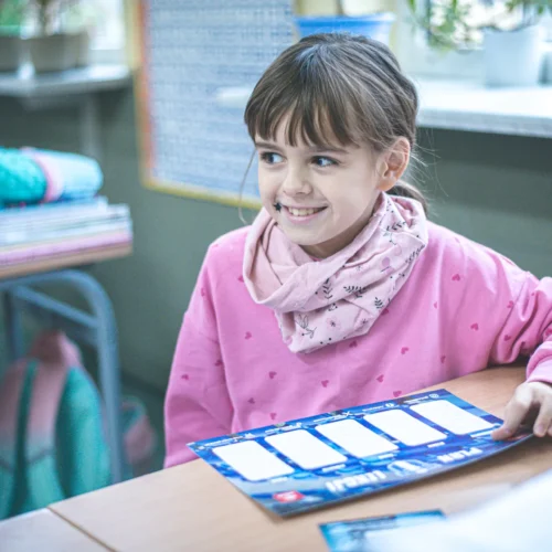 Na zdjęciu widać uśmiechniętą dziewczynkę siedzącą przy szkolnym biurku, która trzyma kartkę z miejscem na wpisy na tle książek i zielonych roślin.