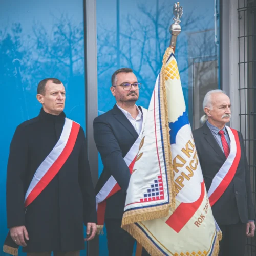 Trzech mężczyzn w formalnych strojach stoi obok siebie, trzymając sztandar, podczas uroczystości na tle niebieskiej ściany.