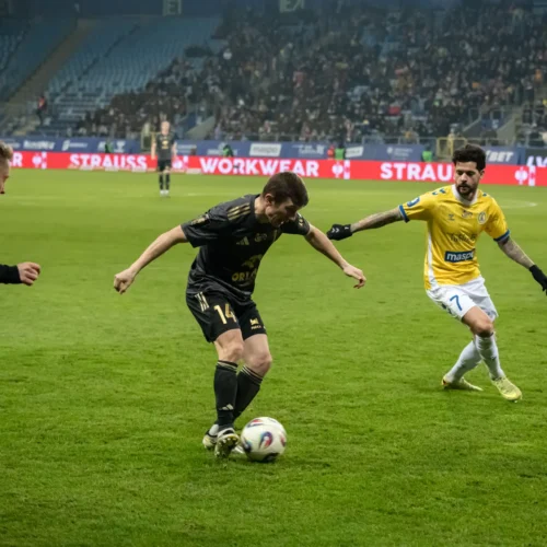 Zdjęcie przedstawia dynamiczną akcję na boisku piłkarskim, gdzie zawodnik w czarnej koszulce stara się przejąć piłkę, podczas gdy dwóch przeciwników zbliża się do niego.