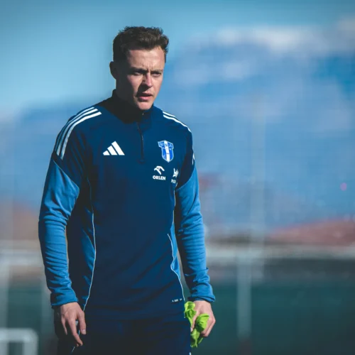 Mężczyzna w niebieskim stroju sportowym stoi na boisku piłkarskim, w tle widać góry i jasne niebo.