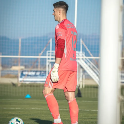 Na zdjęciu widać bramkarza w różowym stroju, który stoi na boisku piłkarskim obok piłki.