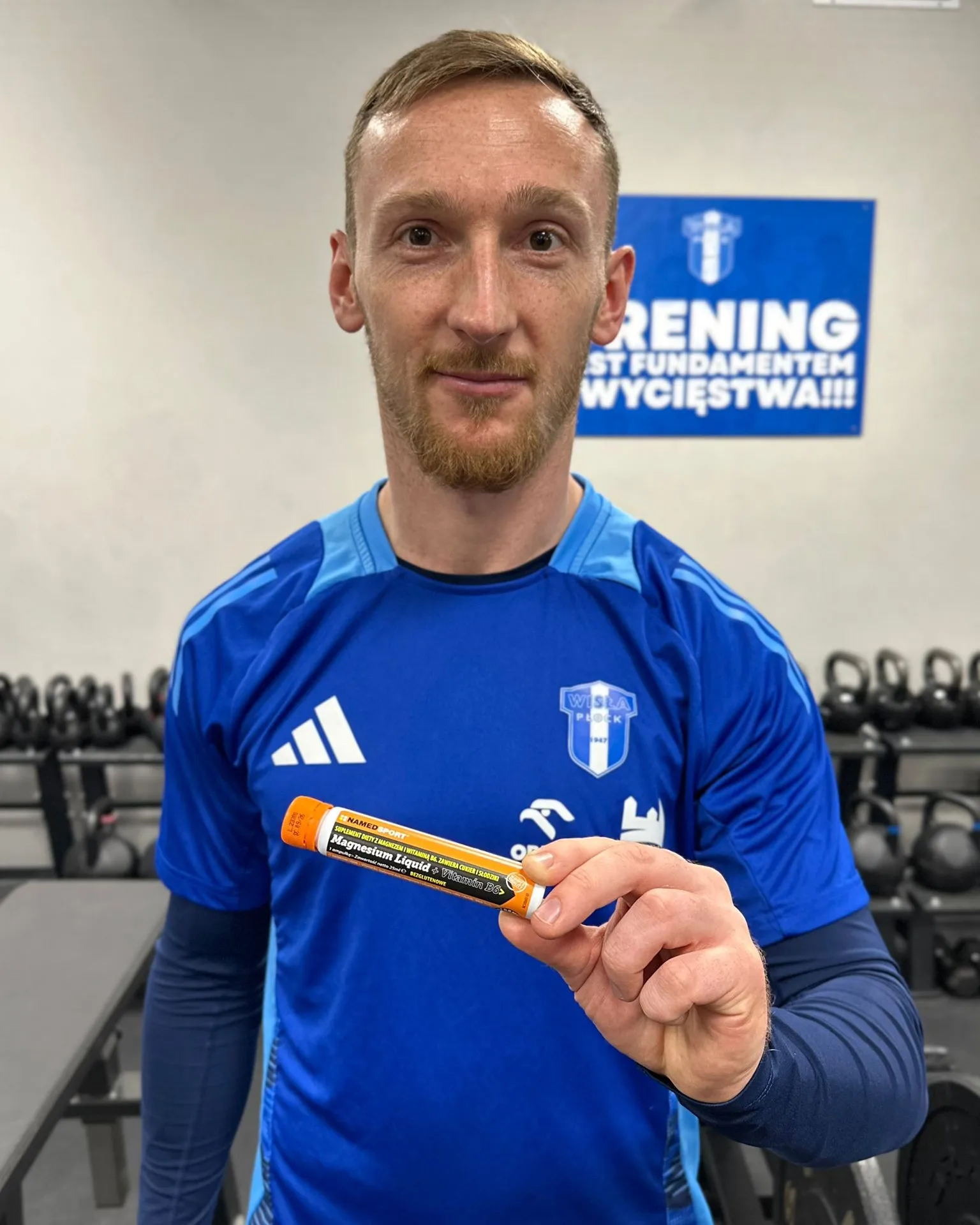 Mężczyzna w niebieskiej koszulce sportowej trzyma butelkę z suplementem diety w siłowni.