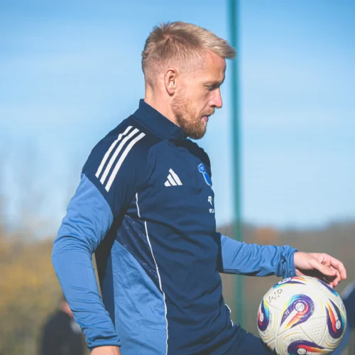 Na zdjęciu widoczny jest mężczyzna w stroju piłkarskim, który ćwiczy z piłką na boisku w słoneczny dzień.