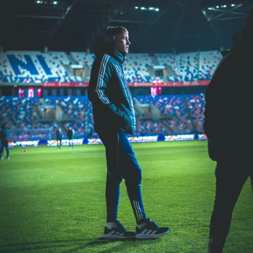 Na zdjęciu widać młodego mężczyznę w sportowej odzieży, stojącego na boisku piłkarskim w ciemności, z widocznym stadionem w tle.