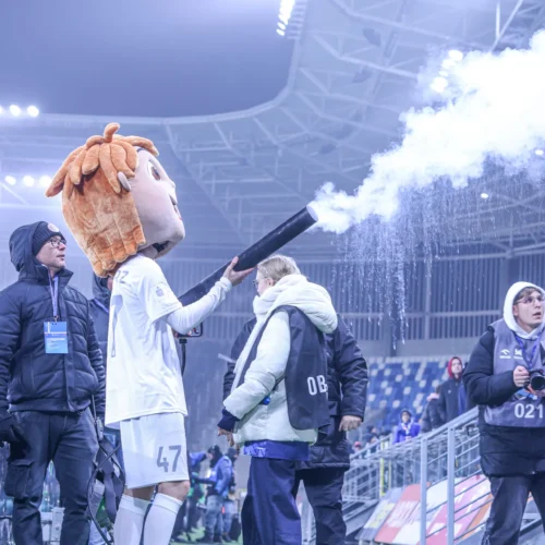 Na zdjęciu przedstawiony jest kostiumowy maskotka z dużą głową, która strzela konfetti z wyrzutni na stadionie, wokół niej znajdują się ludzie w zimowych ubraniach.