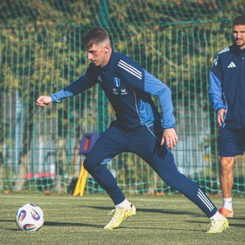 Dwaj piłkarze trenują na boisku, z jednym biegającym z piłką, a drugim obserwującym go w tle.