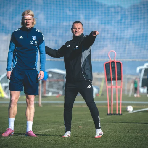 Na zdjęciu widać trenera wskazującego na coś, rozmawiającego z piłkarzem podczas treningu na boisku.