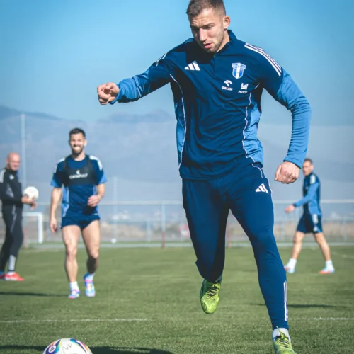 Na zdjęciu widoczny jest piłkarz uczestniczący w treningu na świeżym powietrzu, wykonujący dynamiczny ruch w kierunku piłki.