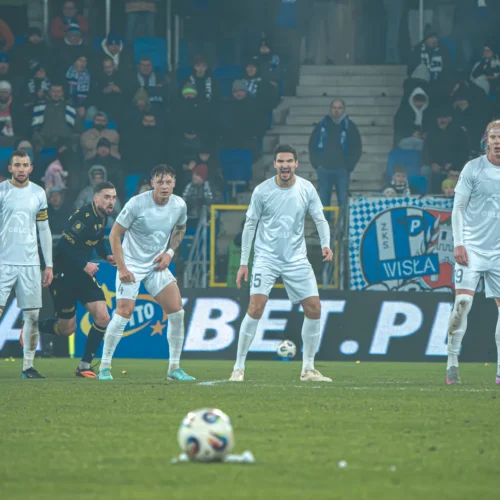 Zawodnicy drużyny piłkarskiej stoją w obronie podczas meczu, przygotowując się na rzut wolny przeciwnika.