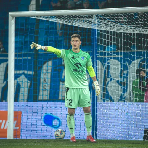 Na zdjęciu widoczny jest bramkarz w zielonym stroju, który wskazuje na swoją drużynę podczas meczu piłkarskiego.