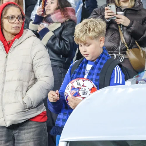 Na zdjęciu widać chłopca trzymającego piłkę oraz kobietę w szarej kurtce, którzy są na stadionie w trakcie meczu.