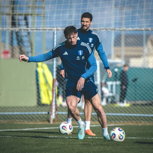 Na zdjęciu dwóch piłkarzy trenuje na boisku, w tle widać siatkę i bramkę do piłki nożnej.