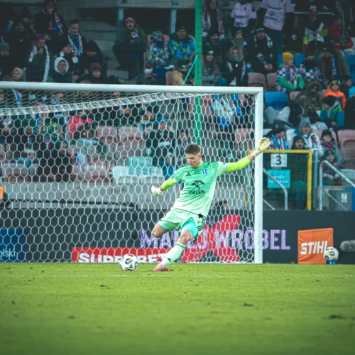 Na zdjęciu widać bramkarza w zielonym stroju, który przygotowuje się do obrony strzału na murawie stadionu, a w tle zgromadzona publiczność.