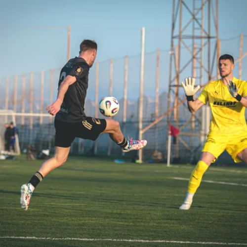 Na zdjęciu widać piłkarza w czarnej koszulce wykonującego strzał do bramki, podczas gdy bramkarz w żółtym stroju stara się obronić uderzenie na boisku.