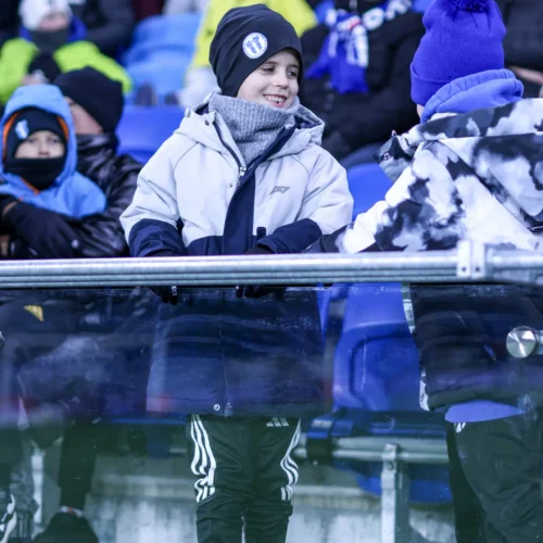 Na zdjęciu widać uśmiechniętego chłopca w zimowej odzieży, który rozmawia z innym dzieckiem na trybunach, w otoczeniu kibiców.