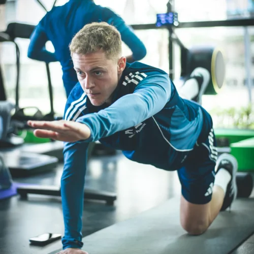 Mężczyzna wykonuje ćwiczenia w siłowni, leżąc na macie i wyciągając rękę do przodu.