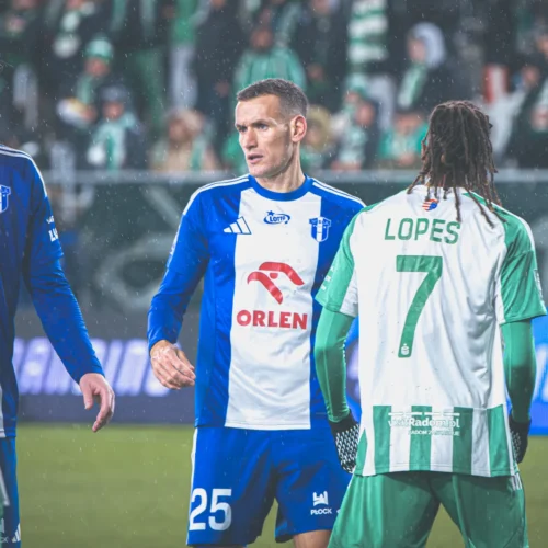 Na zdjęciu widoczna jest scena z meczu piłkarskiego, w której zawodnicy w różnych strojach rozmawiają ze sobą na boisku podczas deszczu.