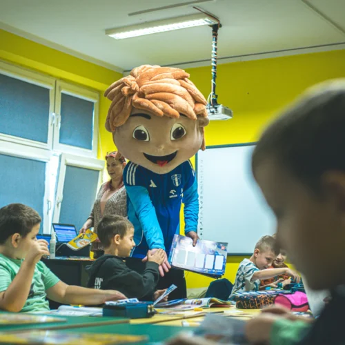 W kolorowej klasie dzieci bawią się podczas wizyty maskotki z dużą, uśmiechniętą głową.