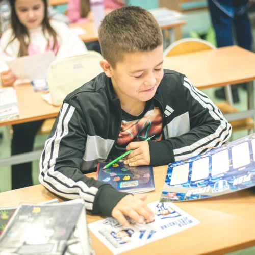 Chłopiec w adidasowej kurtce siedzi przy biurku w klasie, skupiony na rysowaniu i używaniu kolorowych przyborów.