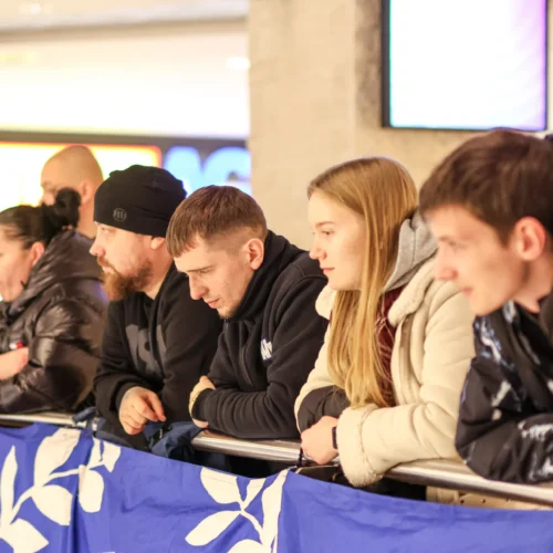 Grupa osób stojących przy barierce, z zauważalnym skupieniem, obserwujących wydarzenie pod dużym ekranem.