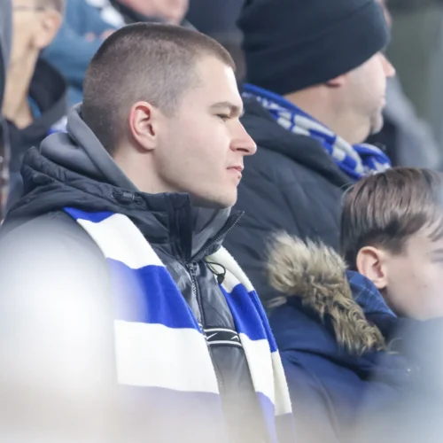 Na zdjęciu widać kibica w szaliku w biało-niebieskie paski, który obserwuje wydarzenia na trybunach stadionu.