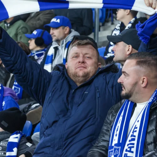Na zdjęciu widać grupę kibiców na trybunach, gdzie jeden mężczyzna trzyma w górze niebiesko-biały szalik, a jego towarzysze również są ubrani w odzież w barwach drużyny.
