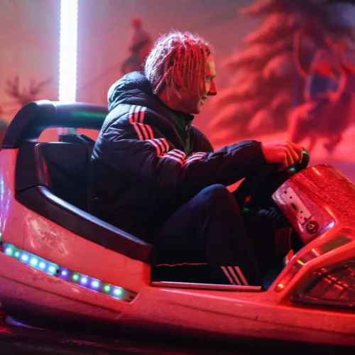 Na zdjęciu widać mężczyznę uśmiechającego się, który siedzi w karuzeli zderzakowej, otoczonej kolorowymi światłami.