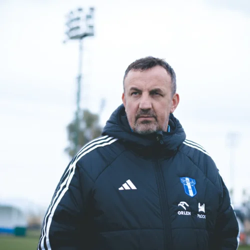 Na zdjęciu widać mężczyznę w czarnej kurtce sportowej z niebieskim logo drużyny, który stoi na stadionie w pochmurny dzień.