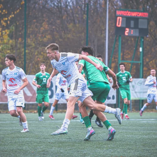Na zdjęciu przedstawiono dynamiczną sytuację na boisku piłkarskim, gdzie zawodnicy w zielonych i białych strojach rywalizują ze sobą, a w tle widoczny jest elektroniczny wynik meczu.