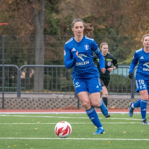 Na zdjęciu widzimy trzy kobiety grające w piłkę nożną na boisku, z jedną zawodniczką w niebieskim stroju biegnącą w kierunku piłki.