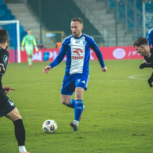 Na zdjęciu widać piłkarza w niebieskiej koszulce, który drybluje z piłką, mijając dwóch rywali w czarnych strojach na boisku.