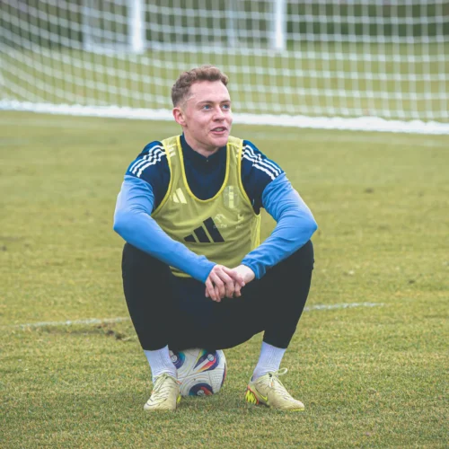 Na zdjęciu widać młodego piłkarza w żółtej kamizelce, siedzącego na murawie boiska i uśmiechającego się, z piłką pod sobą i bramką w tle.