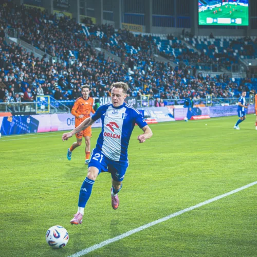 Na zdjęciu widoczny jest piłkarz w niebieskiej koszulce, który biegnie w stronę piłki na stadionie, otoczony przez grupę zawodników i tłum kibiców na trybunach.