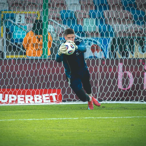 Na zdjęciu widać bramkarza w trakcie obrony strzału piłki na boisku piłkarskim.