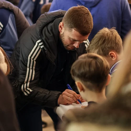 Na zdjęciu widoczny jest mężczyzna w czarnej kurtce, który podpisuje autograf dla dziecka w zatłoczonej przestrzeni.