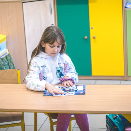 Na zdjęciu siedzi dziewczynka przy biurku w klasie, angażując się w zabawę z kolorową książką.