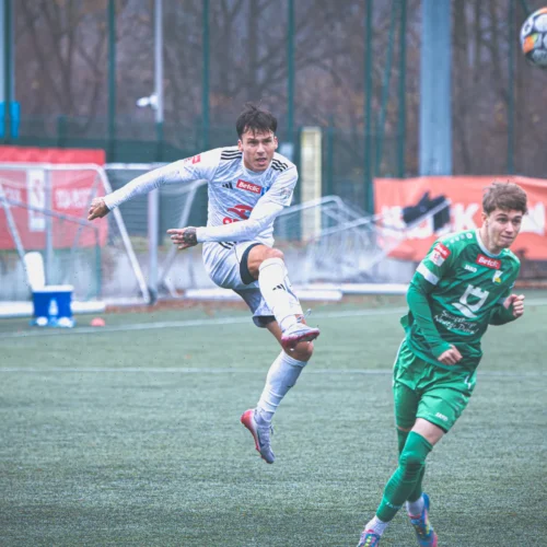 Na zdjęciu dwóch młodych piłkarzy, z których jeden wykonuje spektakularny skok, aby piłką, podczas meczu na boisku trawiastym.