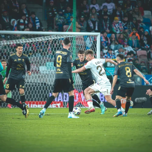 Na zdjęciu widać dynamiczną akcję na boisku piłkarskim, gdzie zawodnik w białym stroju stara się przejść obok kilku przeciwników w czarnych strojach.