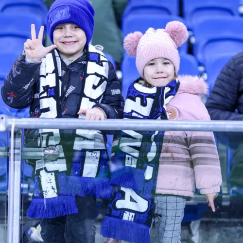 Na zdjęciu widoczni są dwaj mali kibice, chłopiec i dziewczynka, którzy radośnie pozują w szalikach swojej drużyny, na tle stadionowych krzesełek.