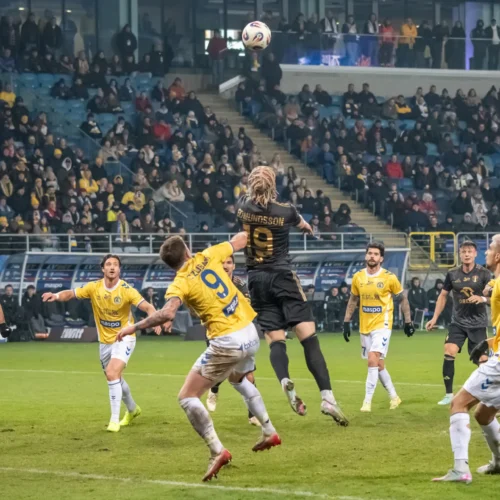 Na zdjęciu widać dynamiczną akcję meczową, w której piłkarz skacze, aby dogonić piłkę, podczas gdy jego przeciwnicy próbują go zablokować.