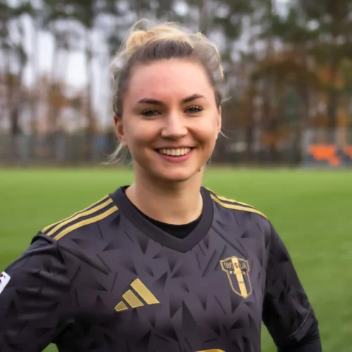 Na zdjęciu uśmiechająca się kobieta w czarnej koszulce piłkarskiej z logo klubu, stojąca na boisku.