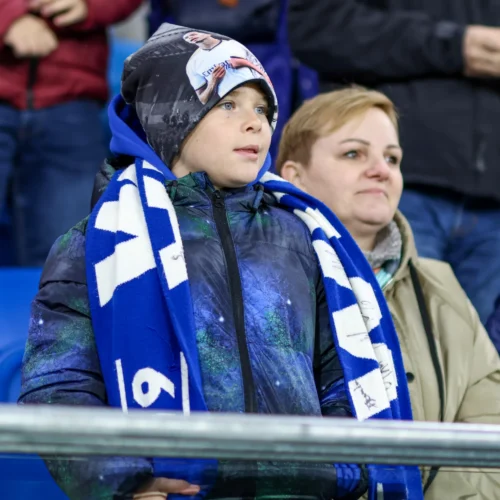 Chłopiec z niebieskim szalikiem, ubrany w kolorową kurtkę, stoi na trybunach wraz z dorosłą osobą, obserwując wydarzenie sportowe.