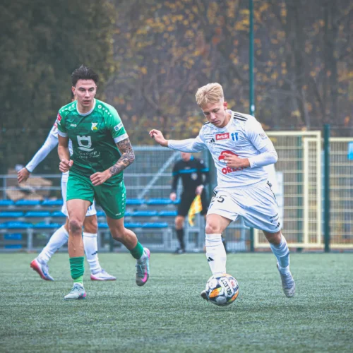 Na zdjęciu dwóch piłkarzy rywalizuje na boisku, z jednym z nich w białej koszulce, a drugim w zielonej, w trakcie meczu piłkarskiego.