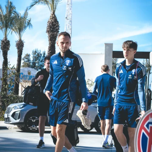 Grupa sportowców w niebieskich strojach treningowych przechodzi obok palm i samochodów w słoneczny dzień.