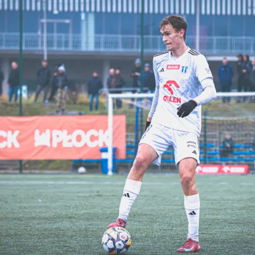 Na zdjęciu zawodnik piłkarski w białym stroju stoi na boisku, trzymając piłkę przy nodze, z widownią w tle.