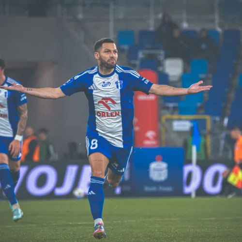 Zawodnik w niebiesko-białej koszulce piłkarskiej świętuje bramkę, wyciągając ramiona w geście radości, podczas meczu na stadionie.