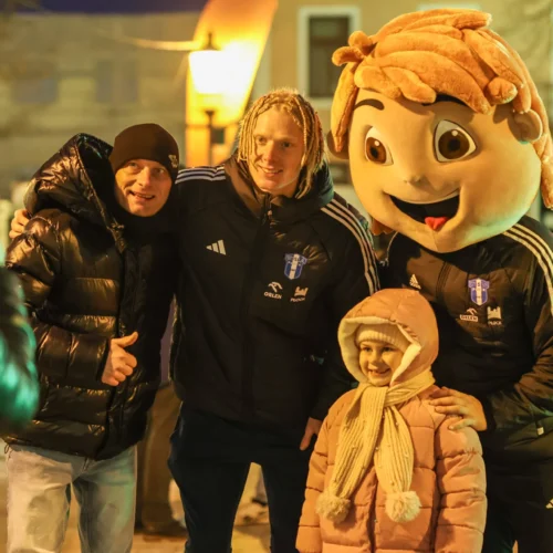 Na zdjęciu grupa osób, w tym dziewczynka w różowej kurtce, pozująca z osobą w kostiumie maskotki oraz dwoma mężczyznami w sportowych kurtkach, w tle widoczne są latarnie i budynki.