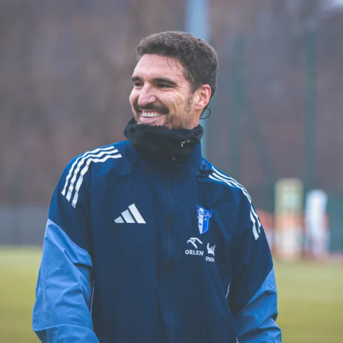 Uśmiechnięty mężczyzna w niebieskiej kurtce sportowej stoi na boisku, ciesząc się z chwili.