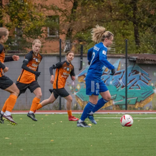 Na zdjęciu widać kobiecą drużynę piłkarską podczas meczu na boisku pokrytym sztuczną trawą.
