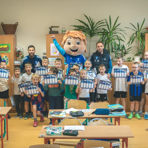 Grupa dzieci w strojach sportowych oraz trenerzy z maskotką cieszą się, trzymając w rękach plany zajęć w klasie szkolnej.